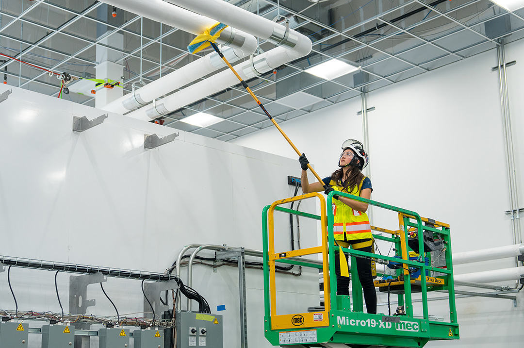 safety-and-security A worker in safety gear, standing on a green lift, cleans overhead pipes with a long-handled tool in an industrial setting, conveying diligence and precision.