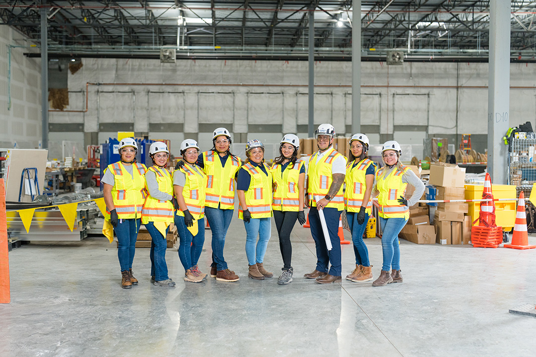 A diverse group of construction workers in hard hats and yellow safety vests stand confidently together in an indoor construction site, smiling.
